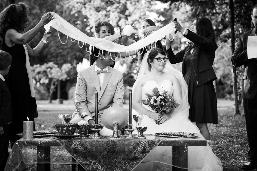 Couple assis devant une table de cérémonie, surplombé par un tissu orné de perles par-dessus lequel une invitée frotte deux morceaux de sucre l'un sur l'autre