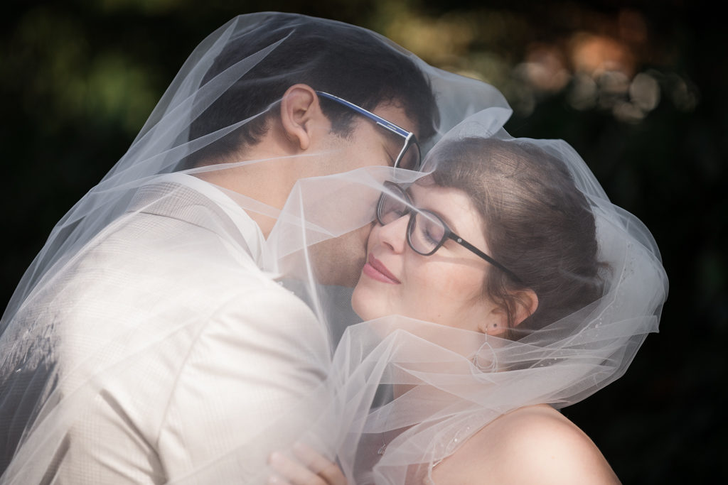 couple sous un voile de mariée, le marié embrassant la mariée qui a les yeux fermés