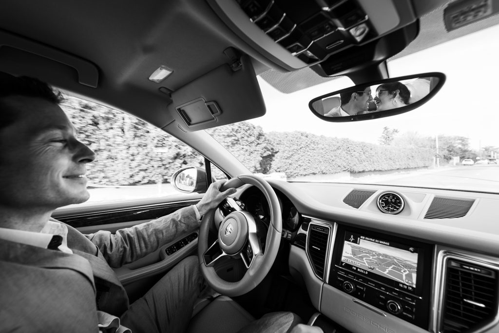 A l'intérieur d'une porsche macane, le conducteur regarde en souriant les mariés s'embrasser dans le rétroviseur central