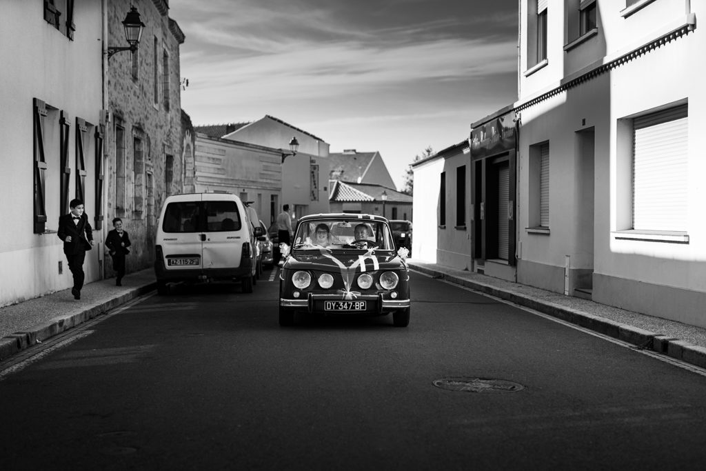 voiture des mariés de face, roulant vers nous, avec sur le trottoir des enfants qui courent en regardant la voiture