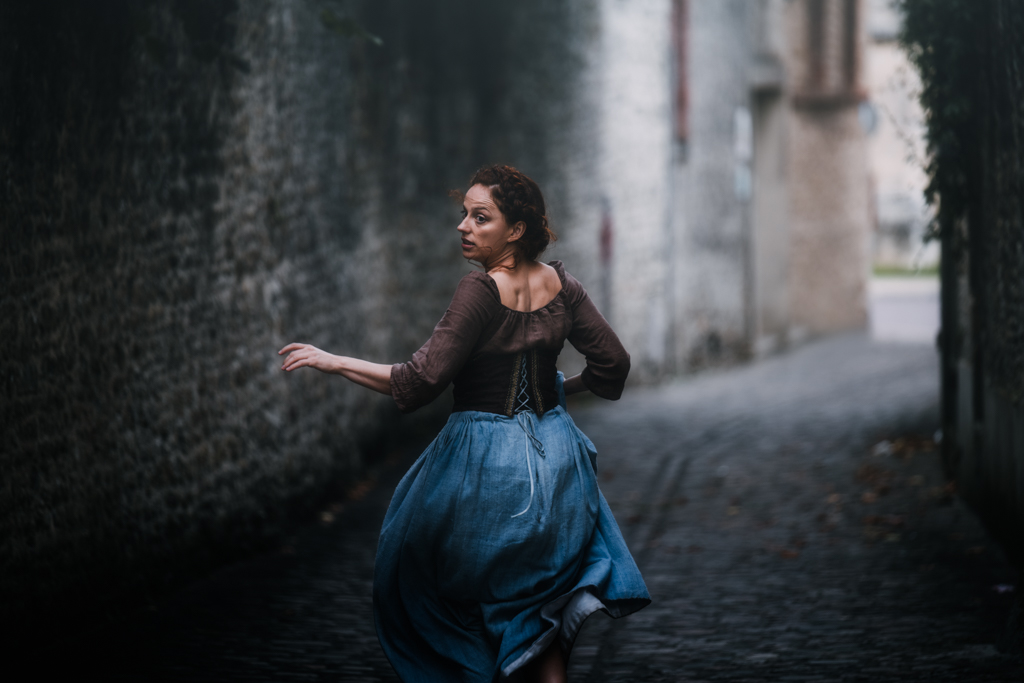 shooting photo en normandie : jeune femme rousse dans une robe d'époque fuyant dans une ruelle