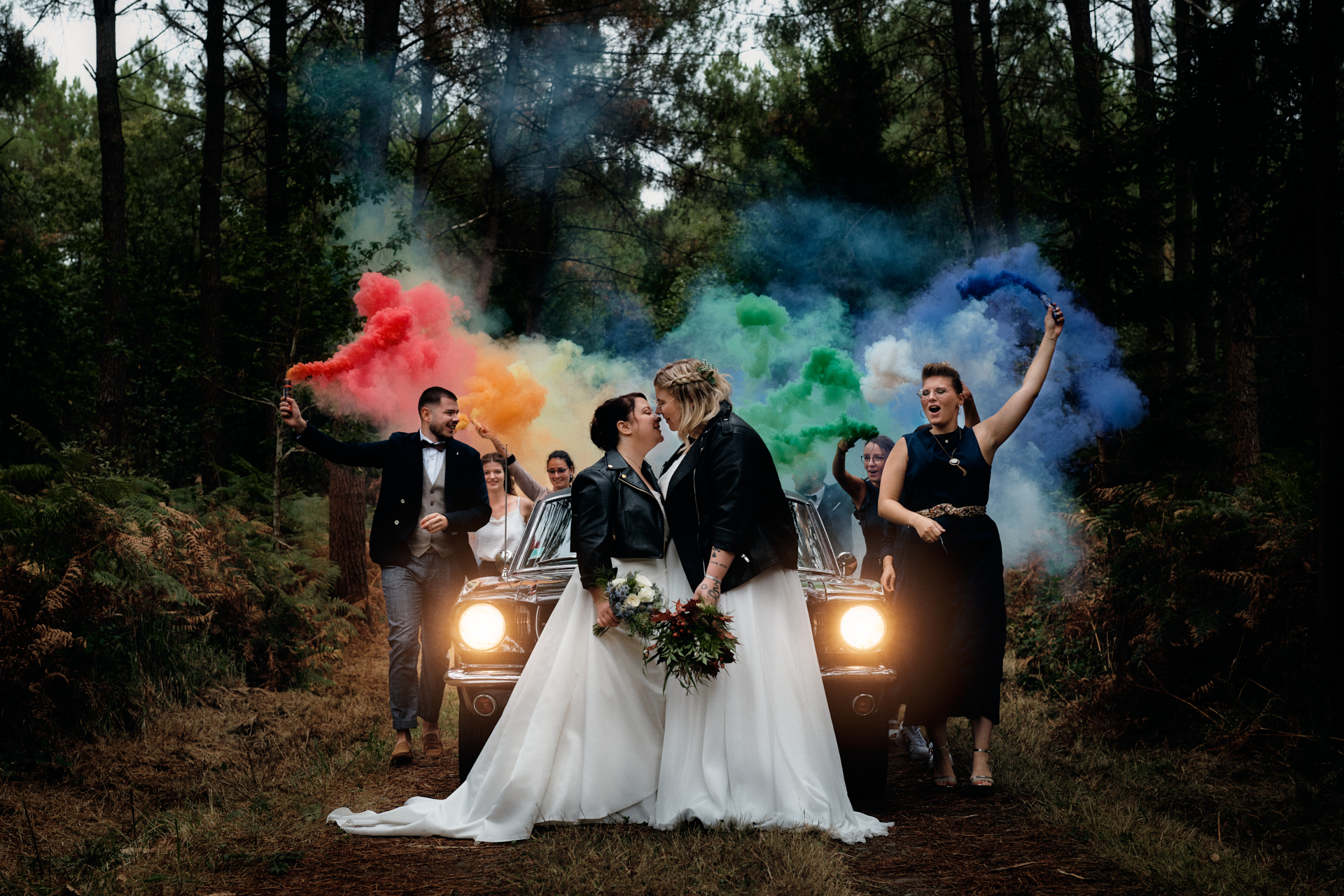 Photo de mariage prise par un photographe de mariage professionnel. Les mariées sont devant leur voiture et leurs témoins arrivent avec des fumigènes formant un arc-en-ciel