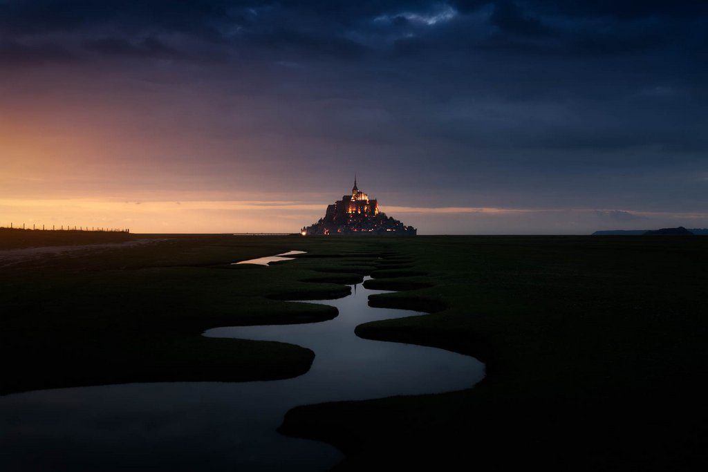Le mont saint michel au coucher de soleil. Des méandres au premier plan dirigent le regard vers le mont, au centre de l'image à l'arrière-plan