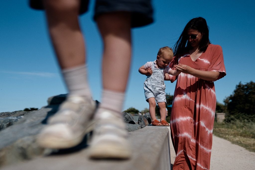 Maman en train d'aider sa fille à marcher sur un muret. Au premier plan on voit les pieds de l'ainé sur le muret
