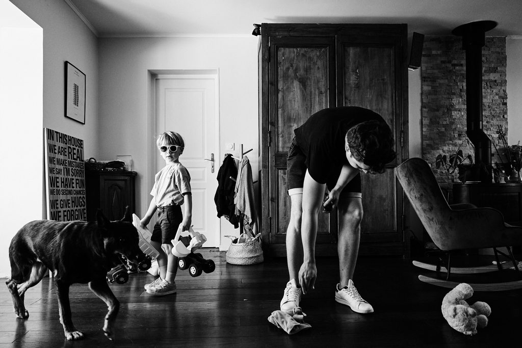 Photo de reportage famille, un enfant marche dans la pièce tandis que le père ramasse les jouets. Le chien passe lui aussi dans le cadre