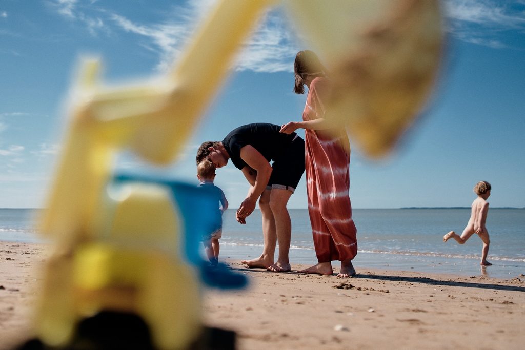 Enfant courant vers la mer pendant que ses parents s'occupent de sa petite soeur. Un jouet au premier plan forme un cadre dans l'image