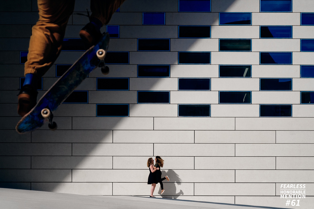Couple de femmes s'embrassant devant un mur avec une ombre projetée triangulaire. Un skateur passe au premier plan en sautant, le skate aligné avec l'ombre