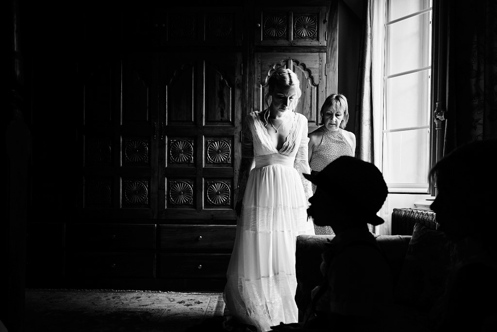 Préparatifs de la mariée lors d'un mariage au chateau de prélong. La mariée vient d'enfiler sa robe aidée de sa maman. La silhouette de son fils assis au premier plan se détache sur le blanc de la robe