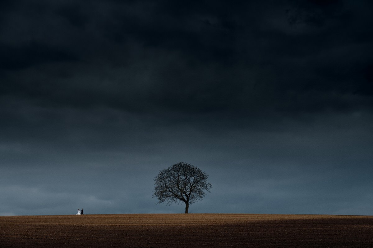 Photo d'un couple au loin sur une colline avec un arbre et un ciel de tempête