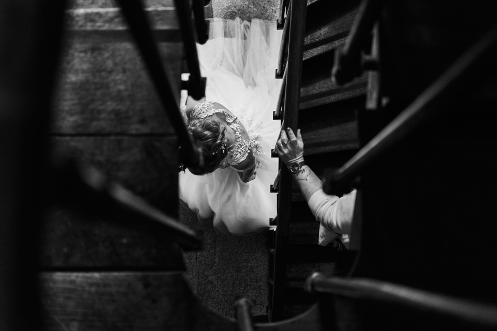Mariée descendant l'escalier, une autre personne la surplombe dont on voit seulement le bras posé sur la rembarde de l'escalier, sur lequel on voit un tatouage du mont saint-michel